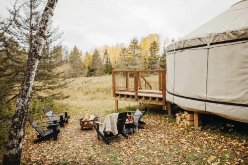 A cozy outdoor scene featuring a yurt, wooden deck, and fire pit surrounded by autumn foliage and trees.