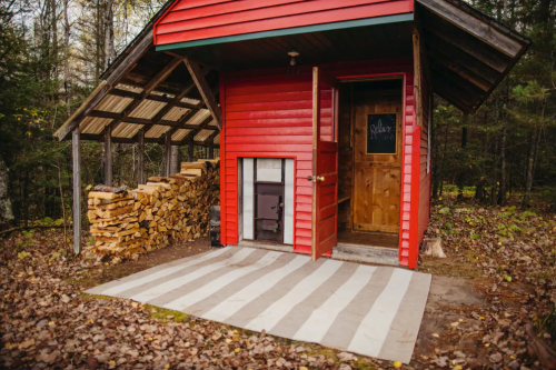 A red cabin with a wooden door, stacked firewood beside it, and a striped mat in front, surrounded by trees.