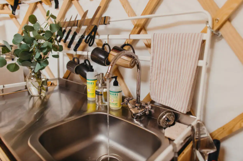 A modern kitchen sink with a faucet, dish soap, and hanging mugs on a wooden wall with kitchen utensils.