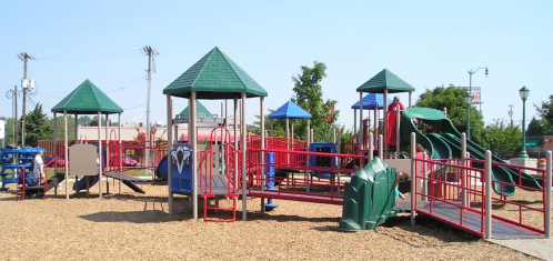 Colorful playground with slides, climbing structures, and shaded areas, surrounded by wood chips and trees.
