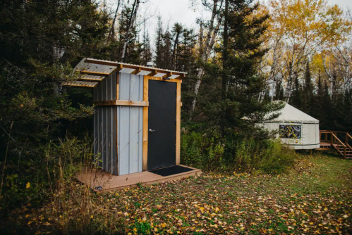 A modern outdoor shower structure beside a yurt, surrounded by trees and autumn foliage.