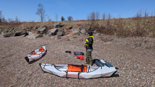 A person in a life jacket stands by two kayaks on a rocky shore, with trees and a clear blue sky in the background.
