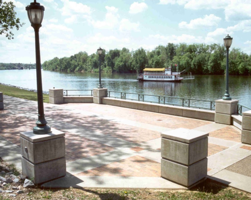 A riverside promenade with lampposts, featuring a boat on the water and lush greenery in the background.