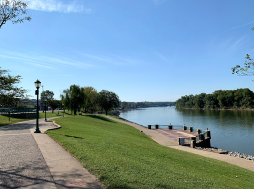 A serene riverside park with a paved path, green grass, and trees under a clear blue sky.