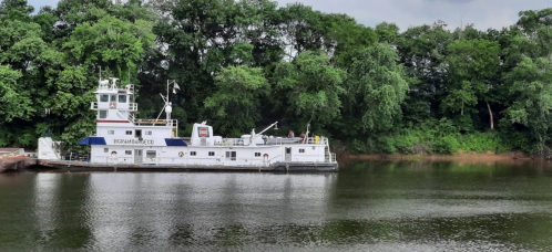 A white boat docked on a calm river, surrounded by lush green trees and a cloudy sky.