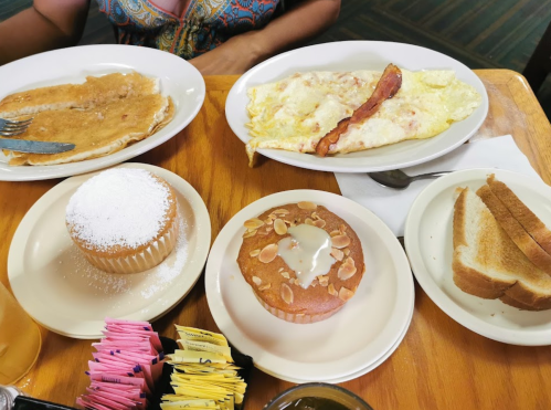 A table with two plates of breakfast: pancakes, an omelet with bacon, and two baked goods, including a cupcake and muffin.