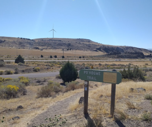 Sign for Po'Hogwe Trail in a dry, open landscape with a wind turbine in the background under a clear blue sky.