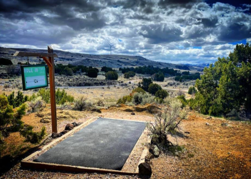 A scenic disc golf tee pad with a sign for Hole 10, surrounded by shrubs and a vast landscape under a cloudy sky.