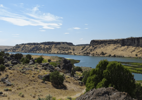 A serene river flows between rocky cliffs and dry land under a clear blue sky. Green vegetation lines the water's edge.