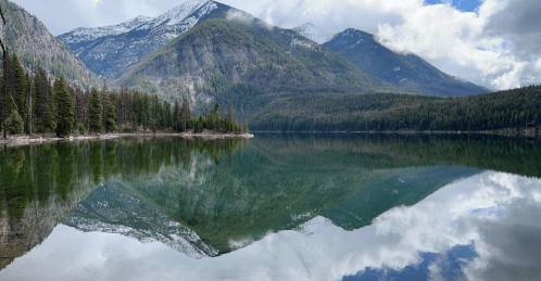 A serene lake reflects mountains and trees under a partly cloudy sky, creating a tranquil natural scene.