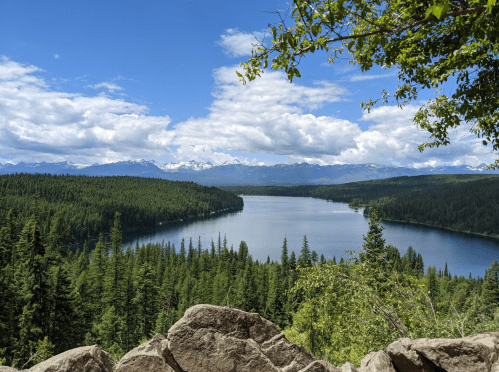 A serene lake surrounded by lush green forests and mountains under a blue sky with fluffy white clouds.