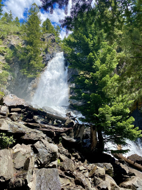 A waterfall cascades down rocky terrain, surrounded by lush green trees and mist rising from the water.