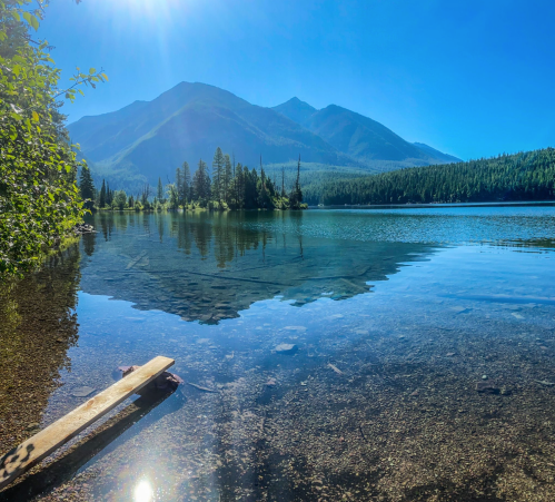 A serene lake surrounded by mountains, with clear water reflecting the blue sky and sunlight.