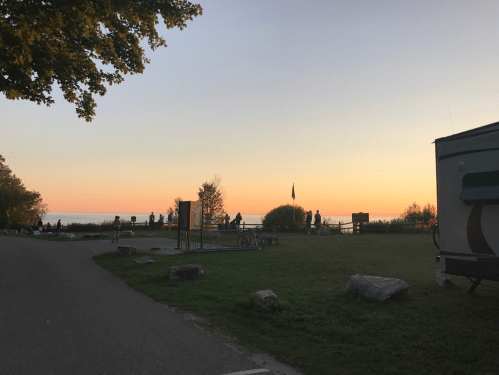 A serene sunset over a lake, with silhouettes of people enjoying the view and a camper parked nearby.