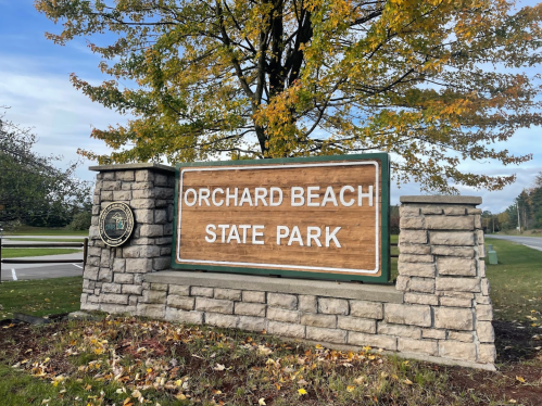 Sign for Orchard Beach State Park, featuring a wooden panel and stone structure, surrounded by autumn foliage.