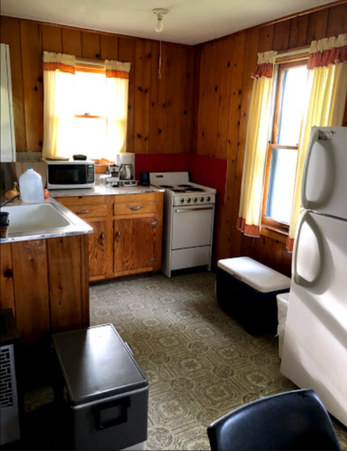 A cozy kitchen with wooden walls, a stove, microwave, and a small table, featuring yellow curtains and patterned flooring.