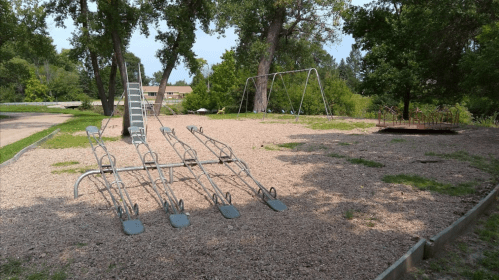 A playground with a slide, swings, and a seesaw, surrounded by trees and gravel.