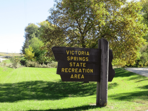 Wooden sign for Victoria Springs State Recreation Area, surrounded by green grass and trees.