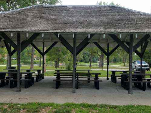 A covered picnic area with wooden tables and benches, surrounded by green grass and trees in a park setting.
