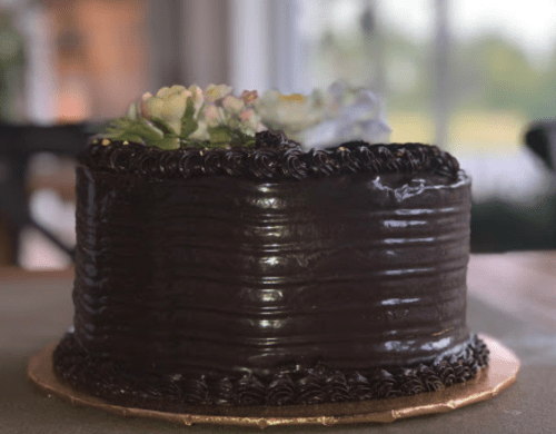 A round chocolate cake with dark frosting and a floral decoration on top, set against a blurred background.