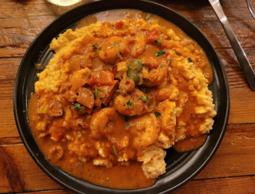 A black plate of shrimp and sauce served over creamy grits, garnished with herbs, on a wooden table.