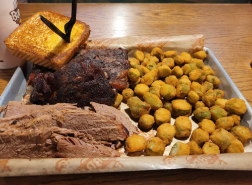 A tray of barbecue featuring sliced brisket, smoked ribs, fried okra, and a piece of toasted bread.