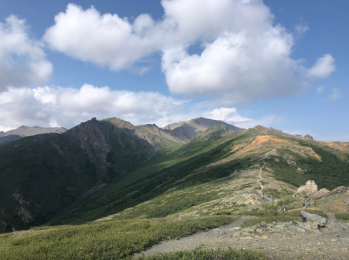 A scenic mountain landscape with rolling hills, green vegetation, and a blue sky dotted with fluffy clouds.