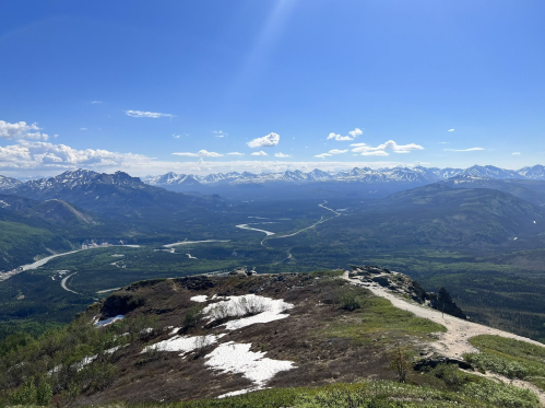 A panoramic view of mountains and valleys under a clear blue sky, with patches of snow on the ground.