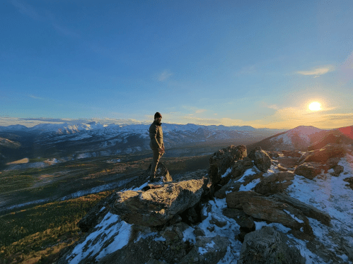 A person stands on a rocky outcrop, overlooking a mountainous landscape at sunset, with snow and trees visible below.