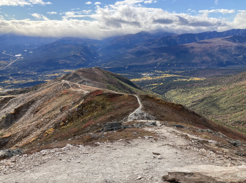 A scenic mountain view with a winding trail, surrounded by valleys and distant peaks under a partly cloudy sky.