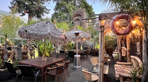 Tropical outdoor dining area with thatched umbrellas, string lights, and lush greenery. Cozy seating arrangements visible.