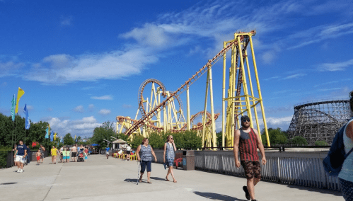 A sunny amusement park scene with a roller coaster, people walking, and colorful flags in the background.
