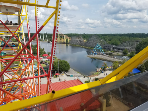 A view from a Ferris wheel overlooking a theme park with roller coasters and a lake under a cloudy sky.