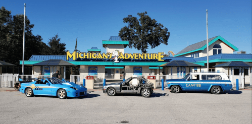 Three blue cars parked in front of the Michigan's Adventure amusement park entrance on a sunny day.