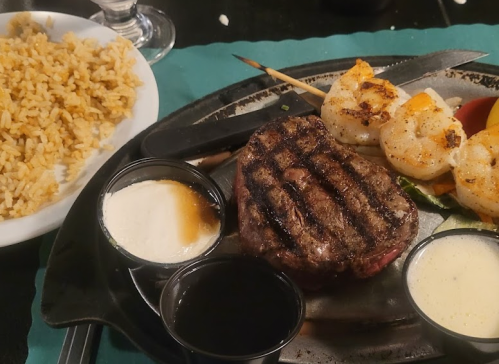 A sizzling plate with grilled steak, shrimp skewers, rice, and dipping sauces.