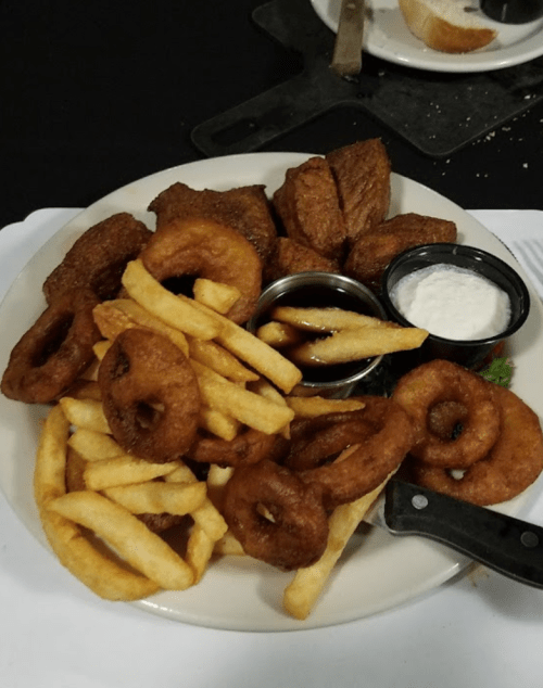 A plate of fried food featuring onion rings, French fries, and two dipping sauces, with pieces of meat on the side.