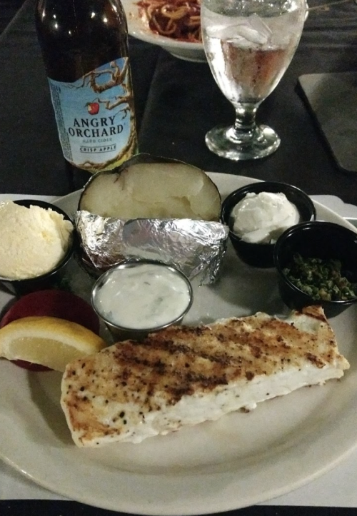 A plate with grilled fish, a baked potato, sour cream, and sauces, alongside a bottle of Angry Orchard cider.
