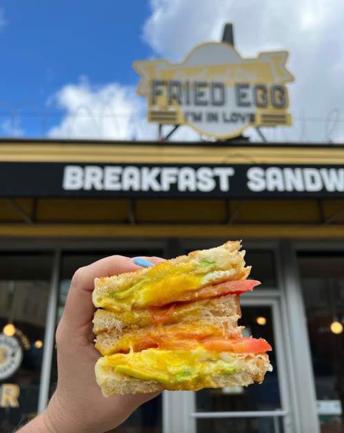 A hand holding a breakfast sandwich with layers of egg, tomato, and green toppings in front of a café sign.