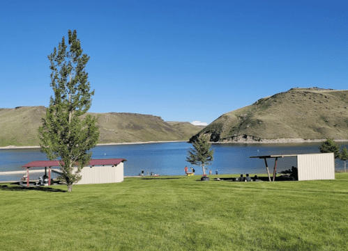 A serene lakeside view with green grass, trees, and picnic shelters against a backdrop of hills and clear blue sky.
