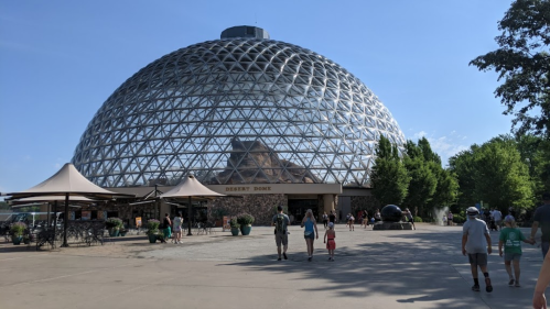 A large glass dome structure surrounded by trees, with people walking towards the entrance on a sunny day.