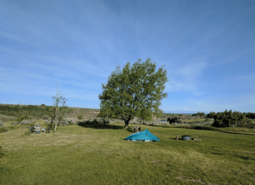 A green field with a large tree and a blue tent under a clear sky, surrounded by distant hills.