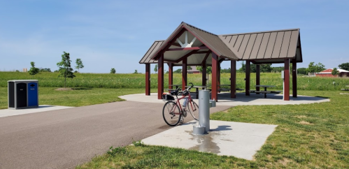 A bike parked near a picnic shelter with a trash can and water fountain, surrounded by green fields and blue sky.
