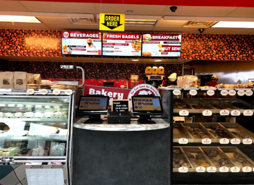 Interior of a bakery with a display case, digital menu boards, and ordering kiosks. Fresh bagels and pastries visible.
