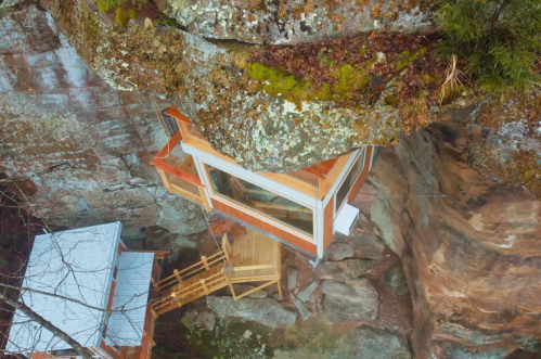 A modern glass cabin perched on a rocky cliff, surrounded by trees and moss, with a wooden deck leading to it.