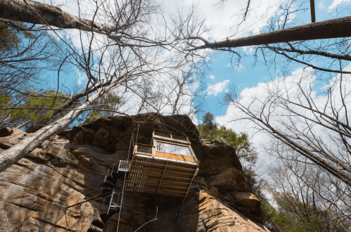 A wooden cabin perched on a rocky cliff, surrounded by bare trees and a bright blue sky with scattered clouds.