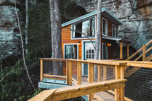 A cozy orange cabin nestled among trees, with a wooden deck and rock formations in the background.