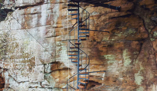 A spiral staircase attached to a rocky cliff, leading upwards against a textured stone background.