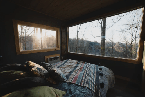 Cozy bedroom with a bed and large windows showcasing a scenic view of trees and hills at sunrise.