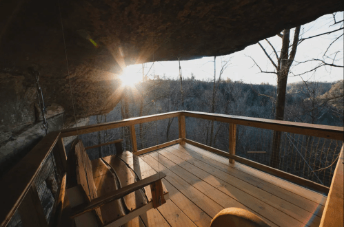 A wooden deck under a rock overhang, with trees and a sunset in the background, creating a serene outdoor space.