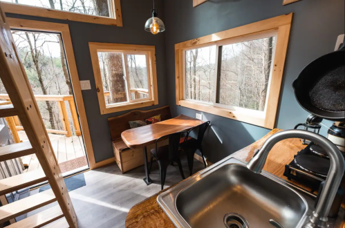 Cozy cabin interior with a wooden table, chairs, and large windows overlooking trees. Modern kitchen sink visible.